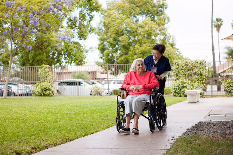 An elderly woman in a wheelchair is being pushed by a caregiver along a paved path in a green outdoor area with trees and bushes. The elderly woman is wearing a bright pink sweater and light gray pants, while the caregiver is dressed in navy blue scrubs and smiling at her.