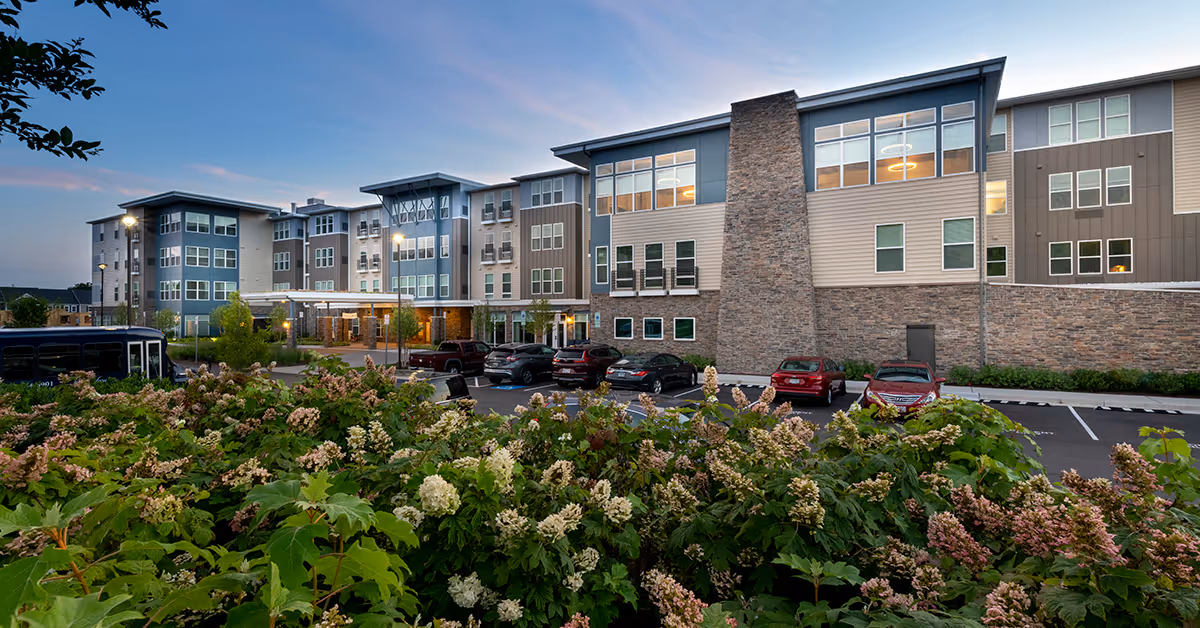 Exterior view of a modern senior living facility building at dusk with multiple floors, large windows, a stone chimney feature, and a parking lot in front. There are flowering bushes in the foreground and several cars parked in the lot.