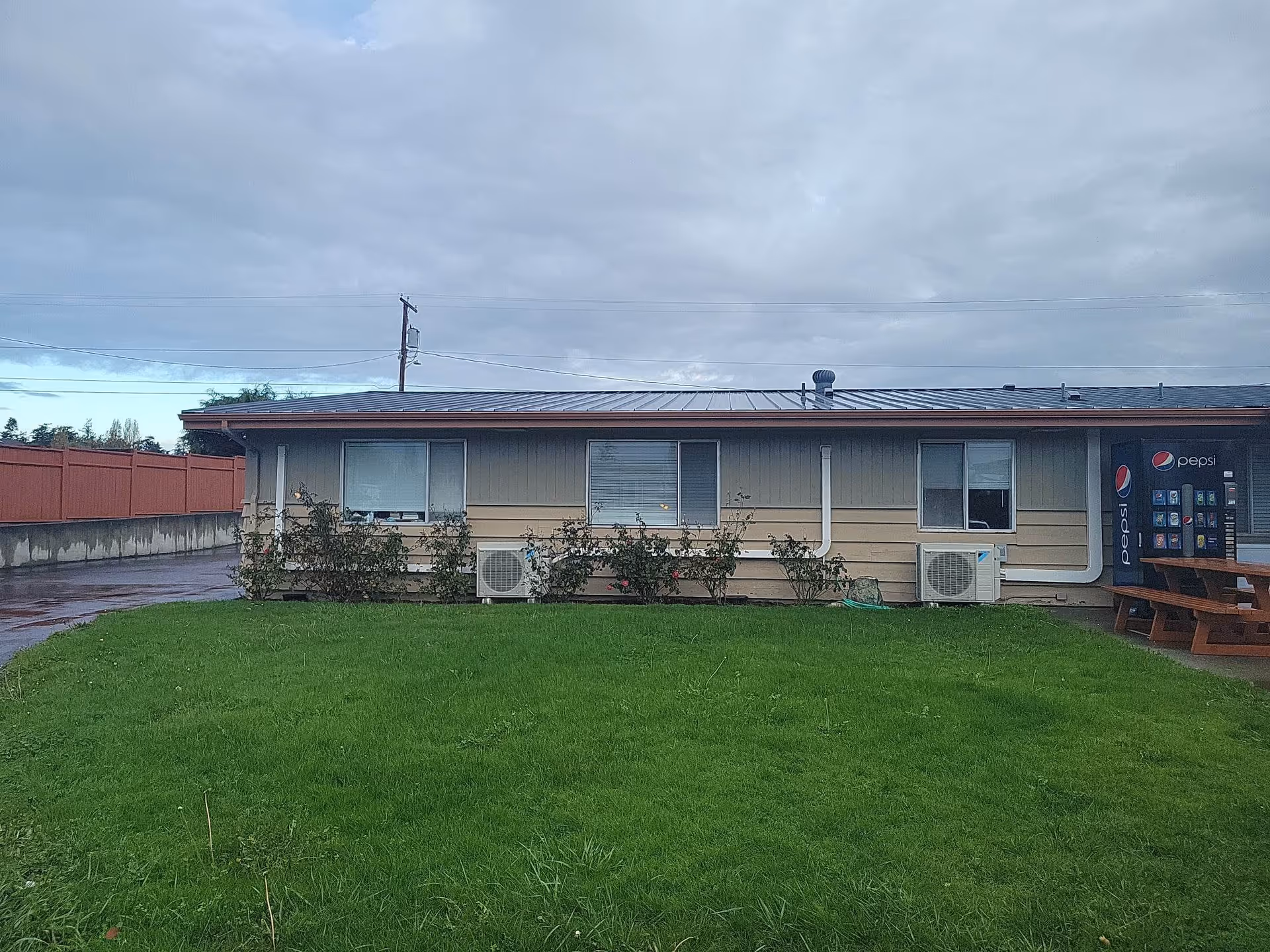 Single-story facility building with a green lawn in front, windows and wall-mounted AC units, and a Pepsi vending machine on the right under a cloudy sky.