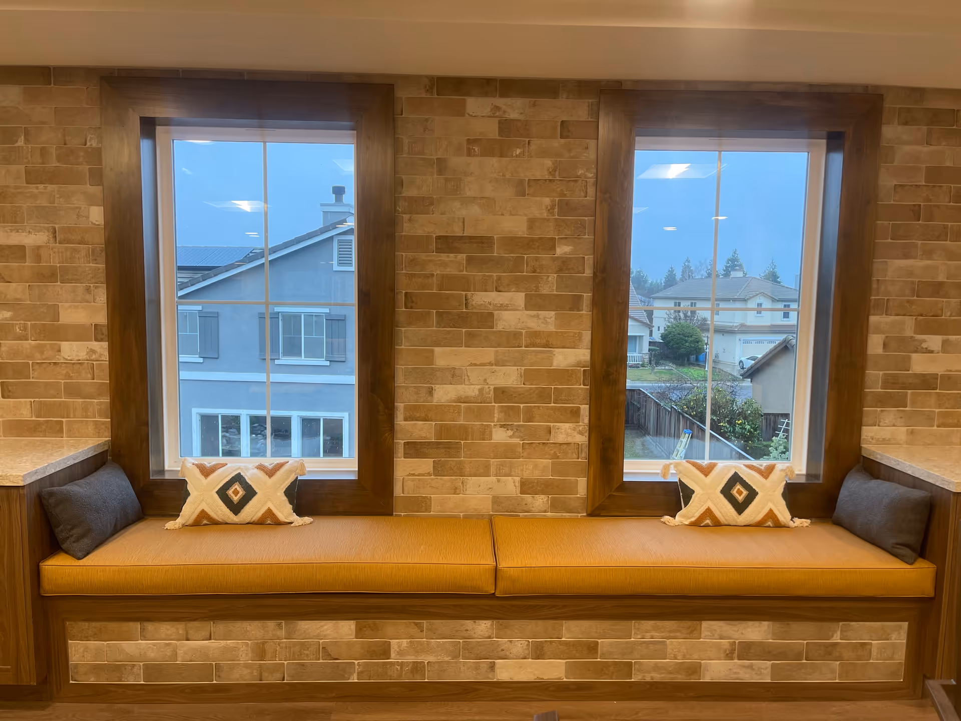 A cozy window seat with mustard yellow cushions and decorative pillows, set against a brick wall with two large windows showing a view of neighboring houses.