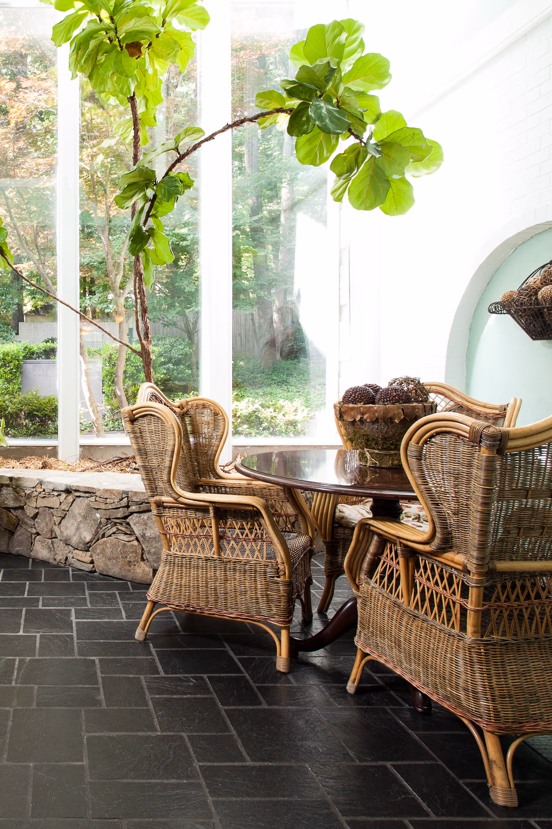 Sunlit sunroom with wicker dining chairs around a round wooden table beside large windows overlooking a garden.