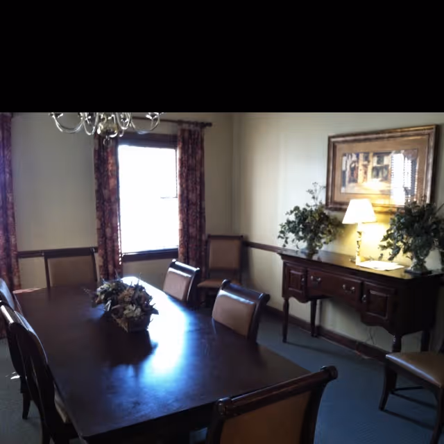 Formal dining room with a long wooden table, upholstered chairs, a sideboard with a lamp and framed artwork, and a curtained window.