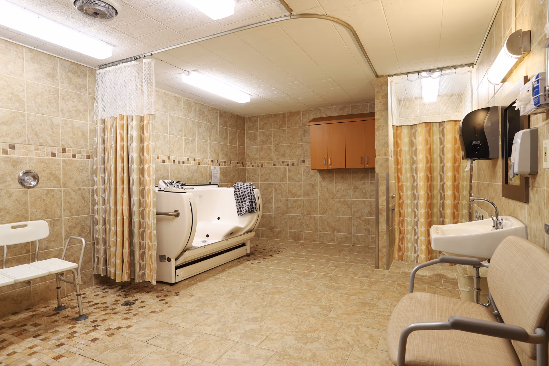 A spacious bathroom with beige tiled walls and floor, featuring a white accessible bathtub with a towel draped over the side, a white shower chair, a sink with a mirror above it, a beige cushioned chair, and two shower curtains with a geometric pattern. There are also wall-mounted cabinets and dispensers for soap and paper towels.