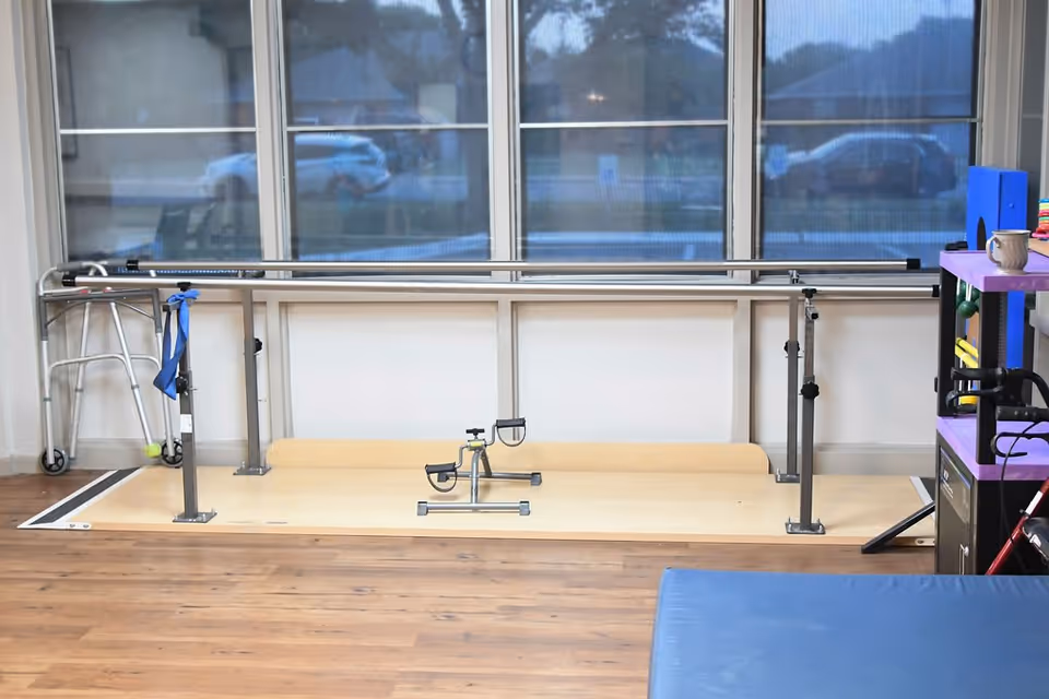 Physical therapy area with parallel bars, a walker, and a pedal exerciser in front of large windows.