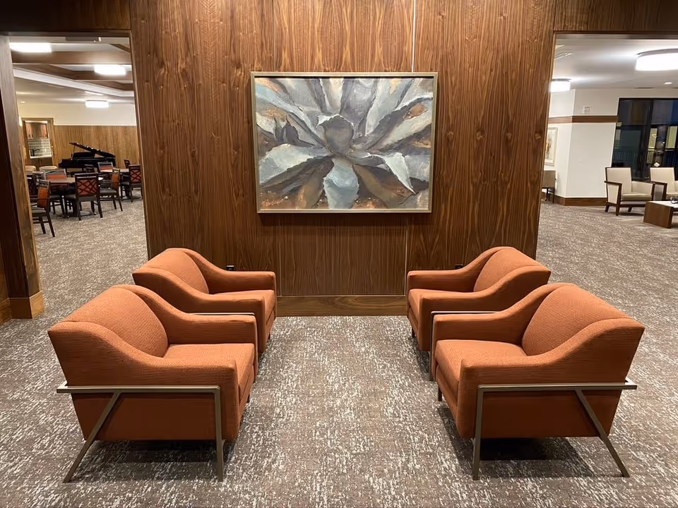 A seating area with four orange upholstered armchairs arranged in two pairs facing each other, set on a patterned carpet. Behind the chairs is a wooden paneled wall with a large abstract painting. In the background, there are additional seating areas and a grand piano visible through an open doorway.