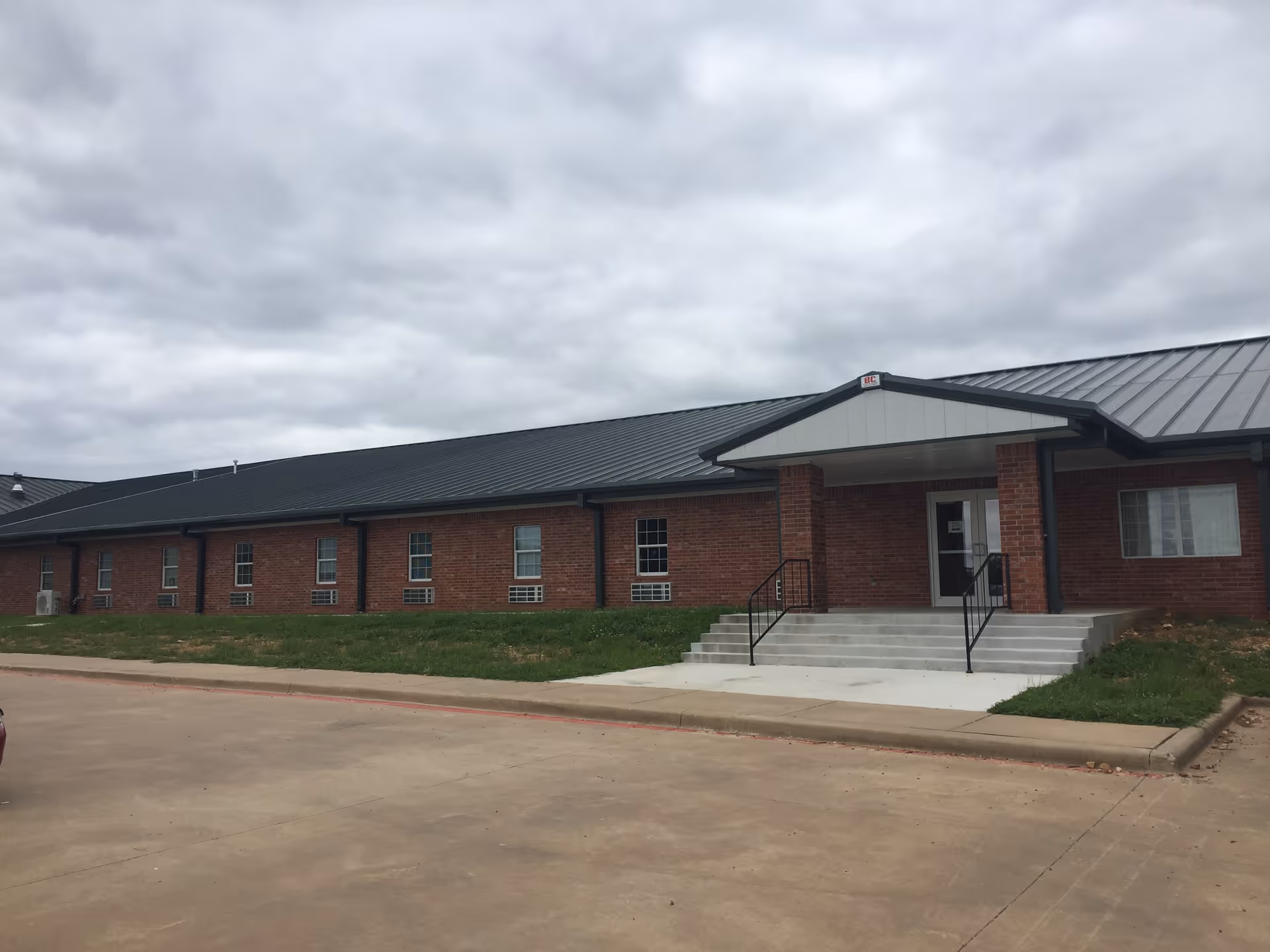 Front exterior of a single-story red brick building with a metal roof and steps leading to the entrance under a cloudy sky.