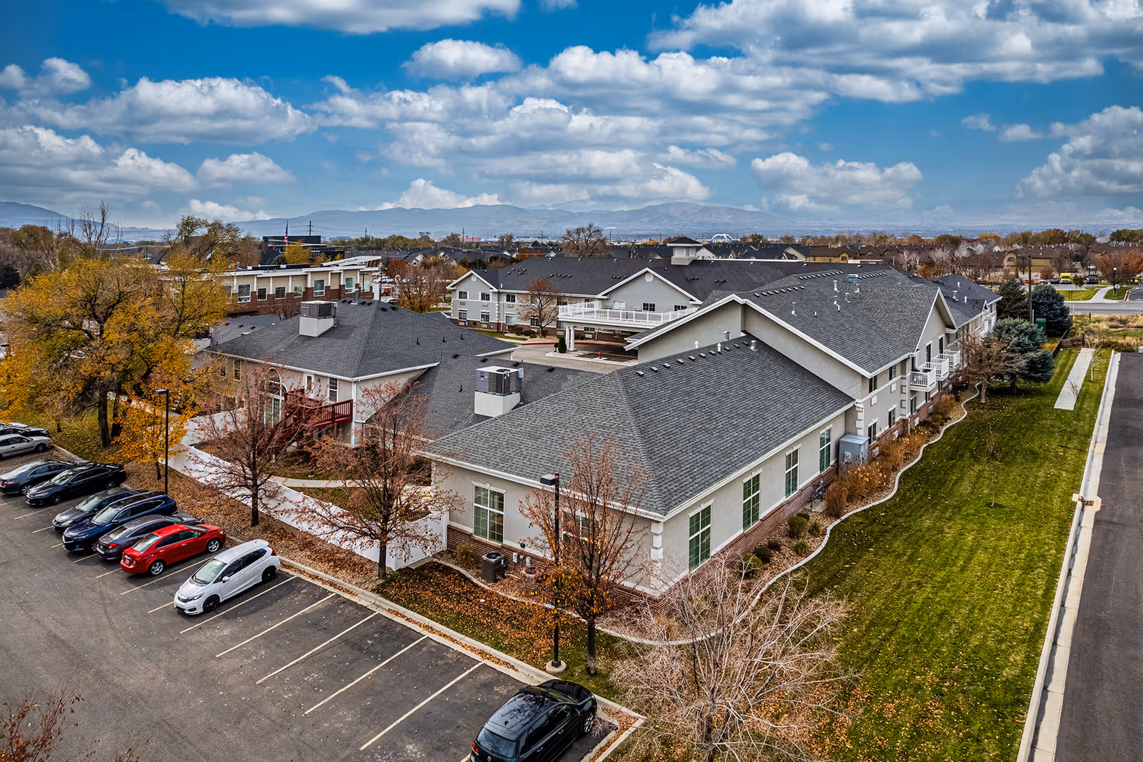 Aerial view of Valencia at Draper senior living facility showing multiple connected buildings with gray roofs, surrounded by trees with autumn foliage, a parking lot with several cars, and a grassy area. Mountains and a partly cloudy sky are visible in the background.