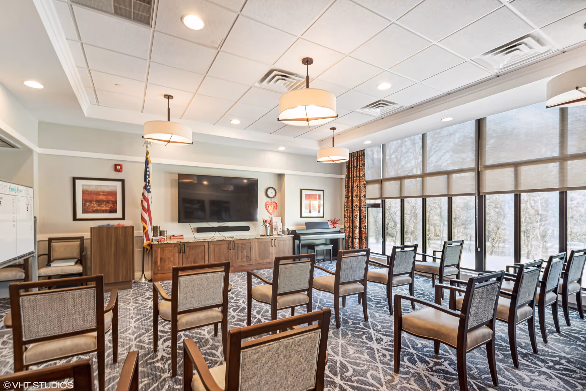 A well-lit meeting or activity room with rows of wooden chairs facing a large flat-screen TV mounted on a wall. The room has large windows with roller shades, patterned carpet, three ceiling lights, an American flag, framed artwork, a piano, and a whiteboard.