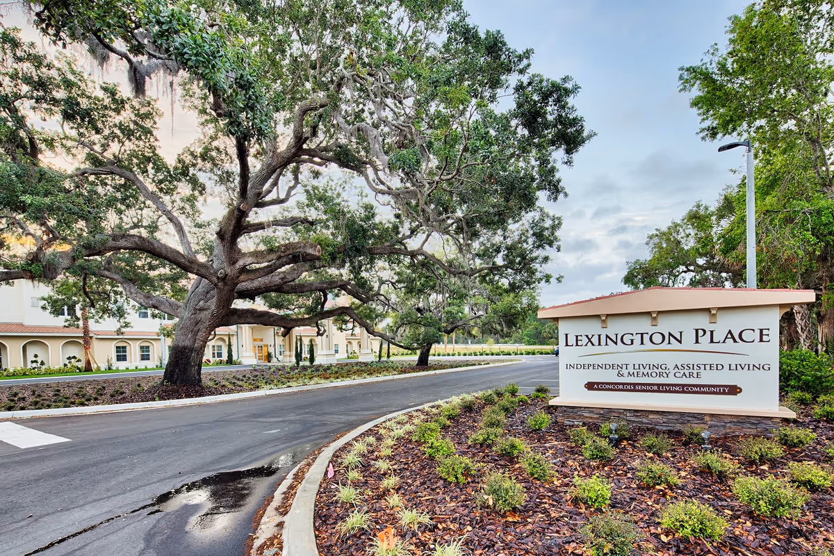 Entrance to Lexington Place senior living community with a large sign displaying the facility name and services, surrounded by landscaped plants and large trees, with the building visible in the background.