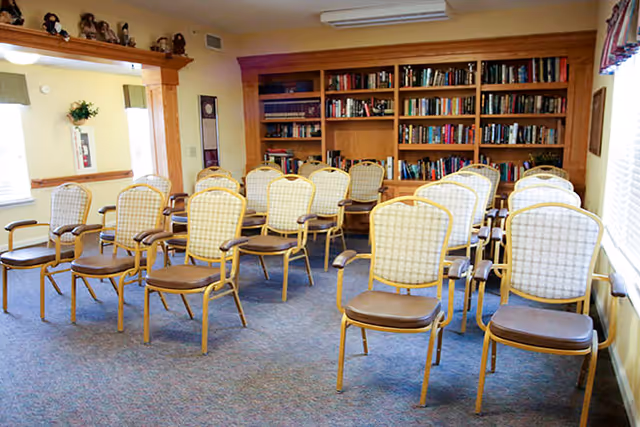 A room with multiple rows of wooden chairs with cushioned seats and backs arranged facing a bookshelf filled with books. The room has carpeted flooring, a window with curtains on the right side, and a wooden framed opening on the left wall.