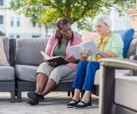 Two elderly women sitting on an outdoor patio sofa, reading books together. One woman has gray hair and is wearing a light green top with a floral scarf, while the other woman has braided hair and is wearing a pink cardigan and beige shorts. There are cushions on the sofa and a tree in the background.