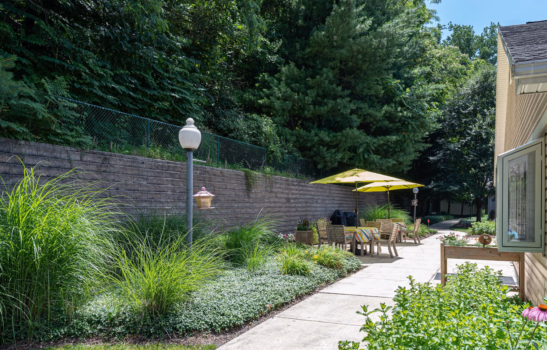 Outdoor patio area with a concrete walkway, surrounded by lush greenery and tall plants. There are several wooden chairs and tables with a yellow umbrella providing shade. A lamppost and a bird feeder are visible near a retaining wall covered with plants. The side of a building with an open window is on the right side of the image.