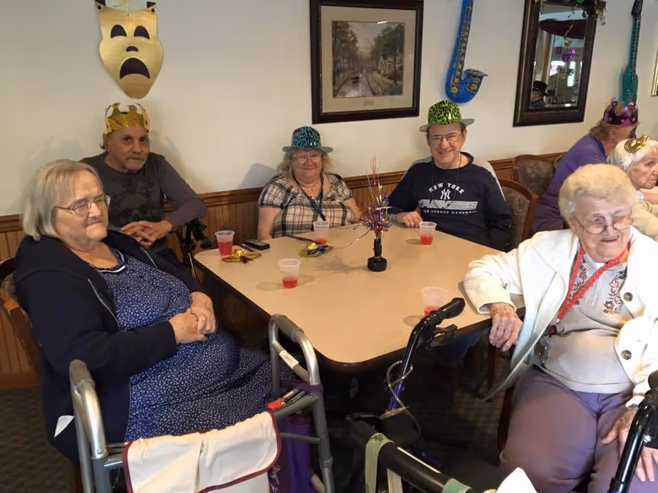 Several elderly residents wearing party hats sit around a table with drinks and a centerpiece in a communal dining/activity room.