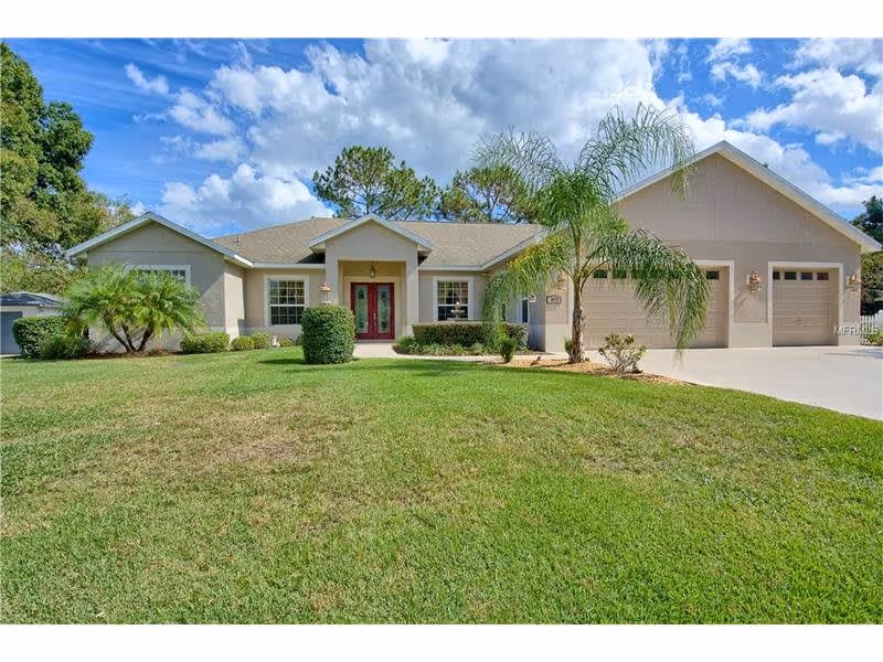 Single-story residential building with a well-maintained front lawn, palm trees, and a driveway leading to a two-car garage under a partly cloudy blue sky.