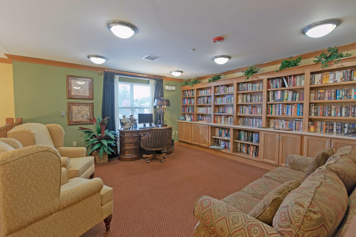 A cozy library room with a large wooden bookshelf filled with books along one wall. There is a patterned sofa on the right and two beige armchairs on the left. A wooden desk with a computer and decorative items is positioned in front of a window with dark curtains. The room has green and yellow walls, carpeted floor, and ceiling lights.