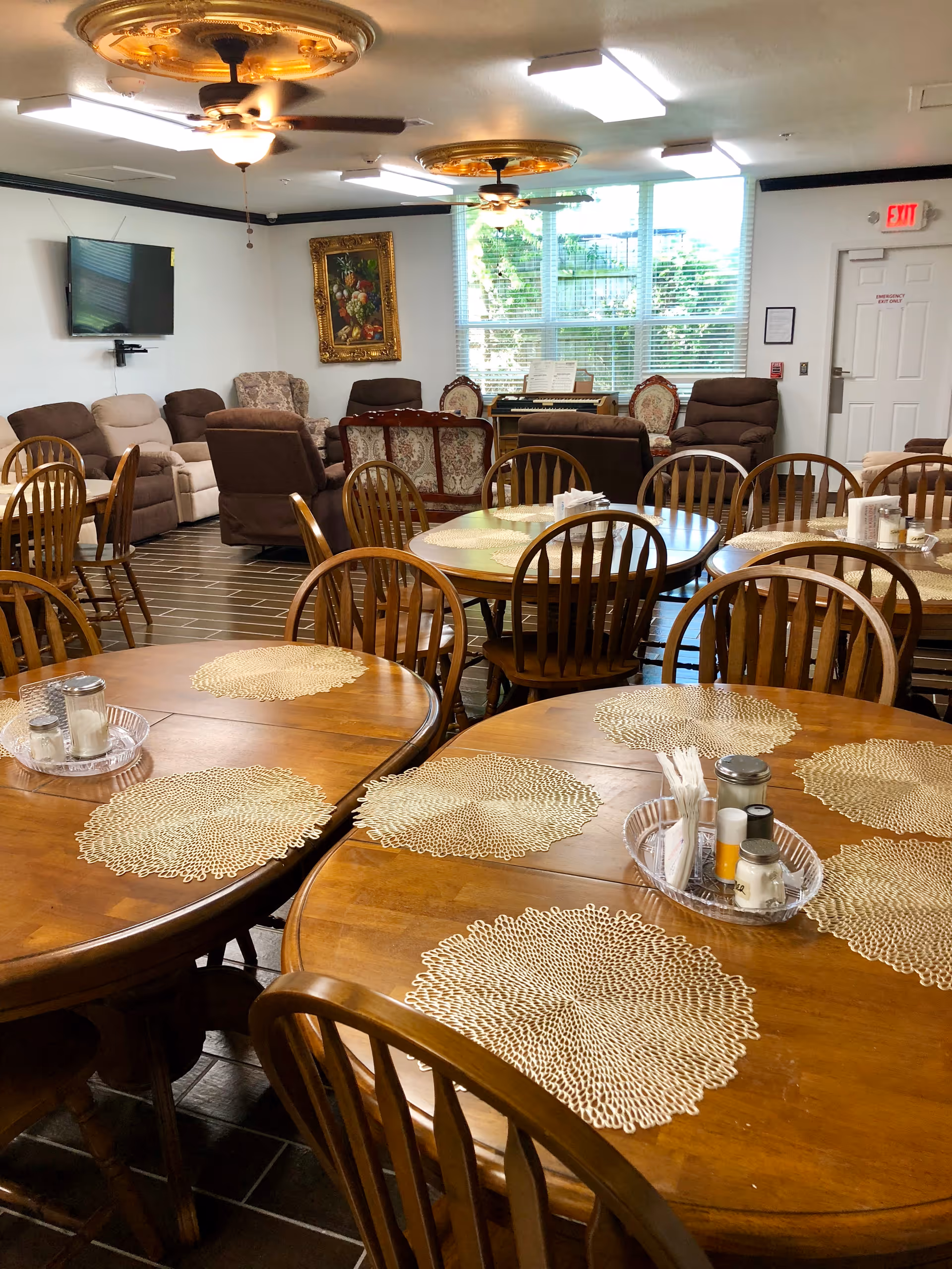 A dining room in an assisted living facility with several round wooden tables and matching wooden chairs. Each table has decorative placemats and condiment trays. In the background, there is a seating area with various upholstered armchairs and a wall-mounted TV. Large windows let in natural light, and ceiling fans with lights hang from the ceiling.