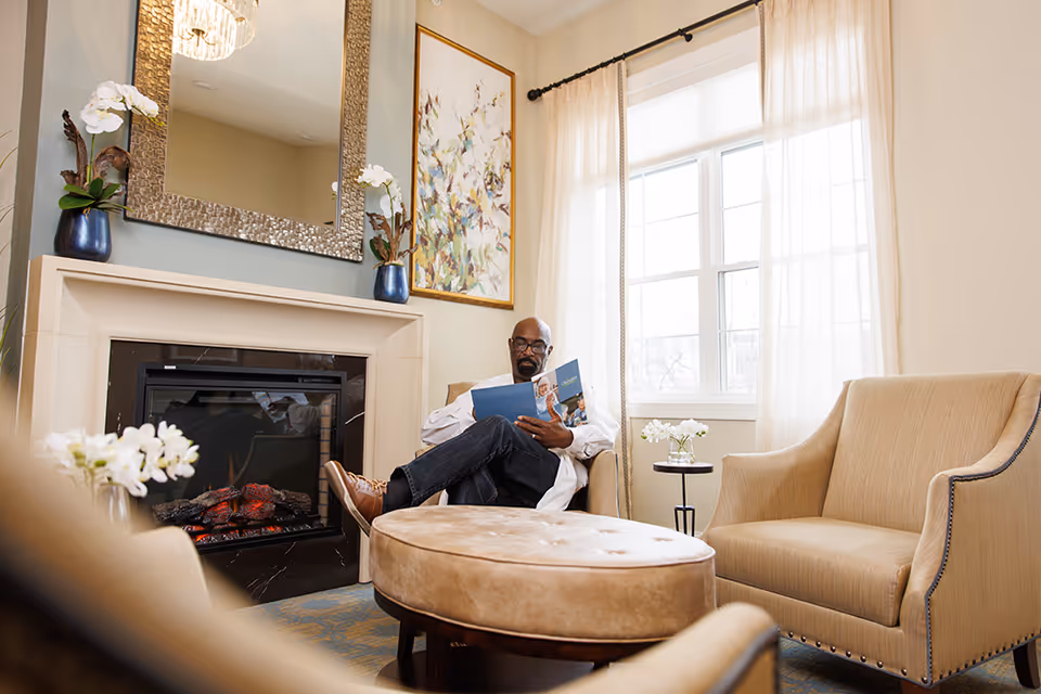 A person sits reading in a bright lounge with armchairs, an ottoman, a fireplace, and a large window.