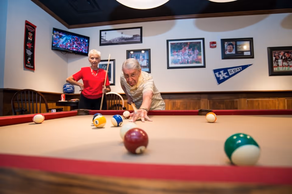 An elderly man and woman playing pool in a recreational room. The man is aiming to strike the cue ball with a pool cue while the woman stands behind him holding her cue stick. The room has wood paneling, framed sports photos, a Kentucky pennant on the wall, and a television showing a sports game.