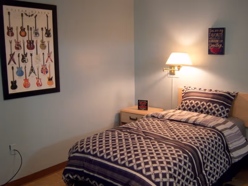 A single bed with a patterned bedspread and pillow in shades of purple and white is positioned against a light-colored wall. Above the bed is a wall-mounted lamp emitting a warm light. To the right of the bed is a small wooden nightstand with a small decorative item on it. On the wall above the nightstand is a small framed sign with a sentimental quote. On the left wall, there is a large framed poster titled 'Guitar Heaven' displaying various guitar illustrations.