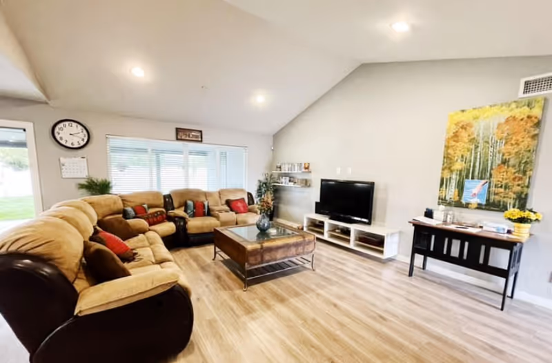 Bright senior living lounge with beige sectional, coffee table, TV, and wall art.