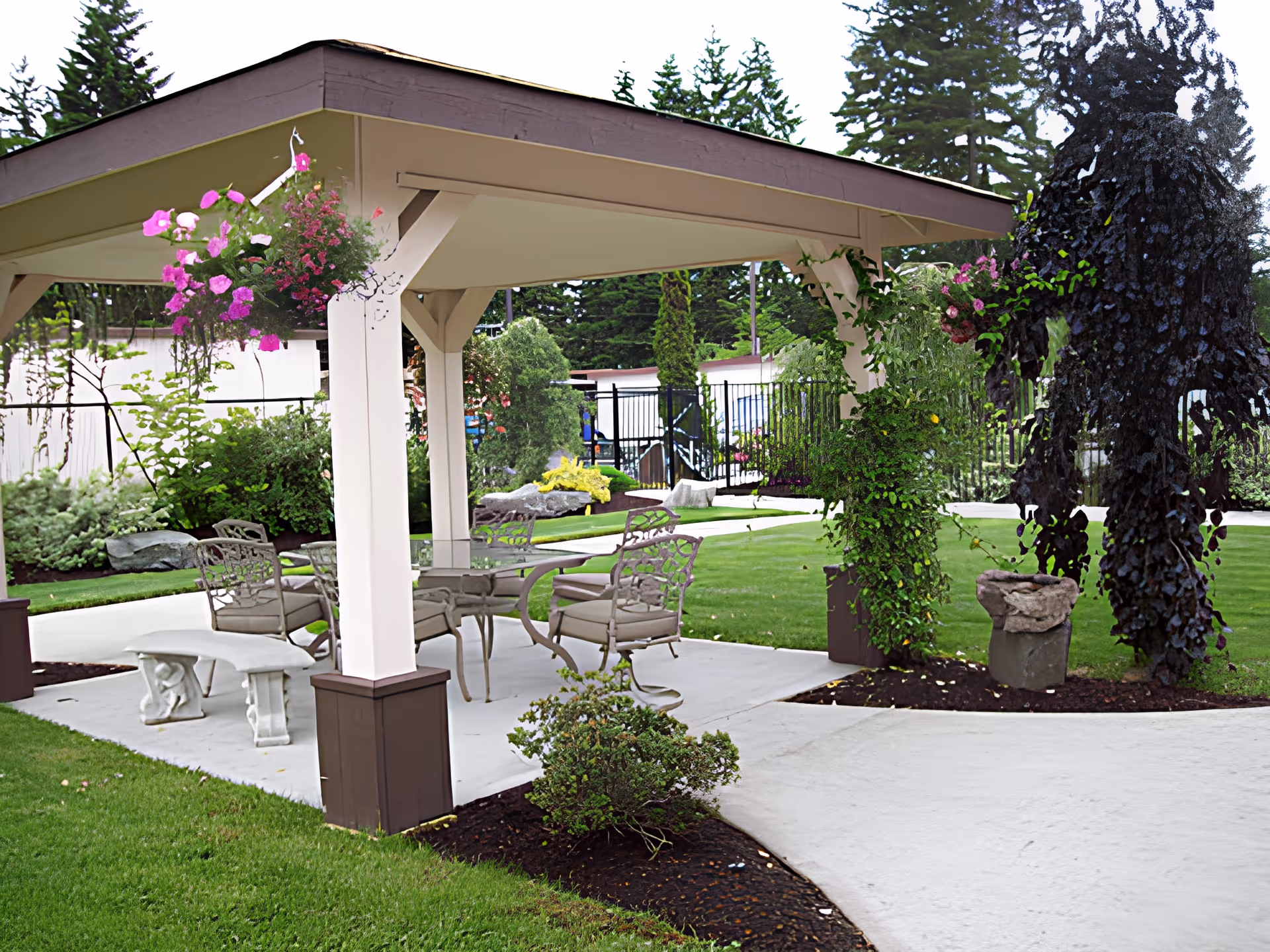 Covered outdoor seating area with metal chairs and a table under a gazebo in a landscaped garden with hanging flowers.