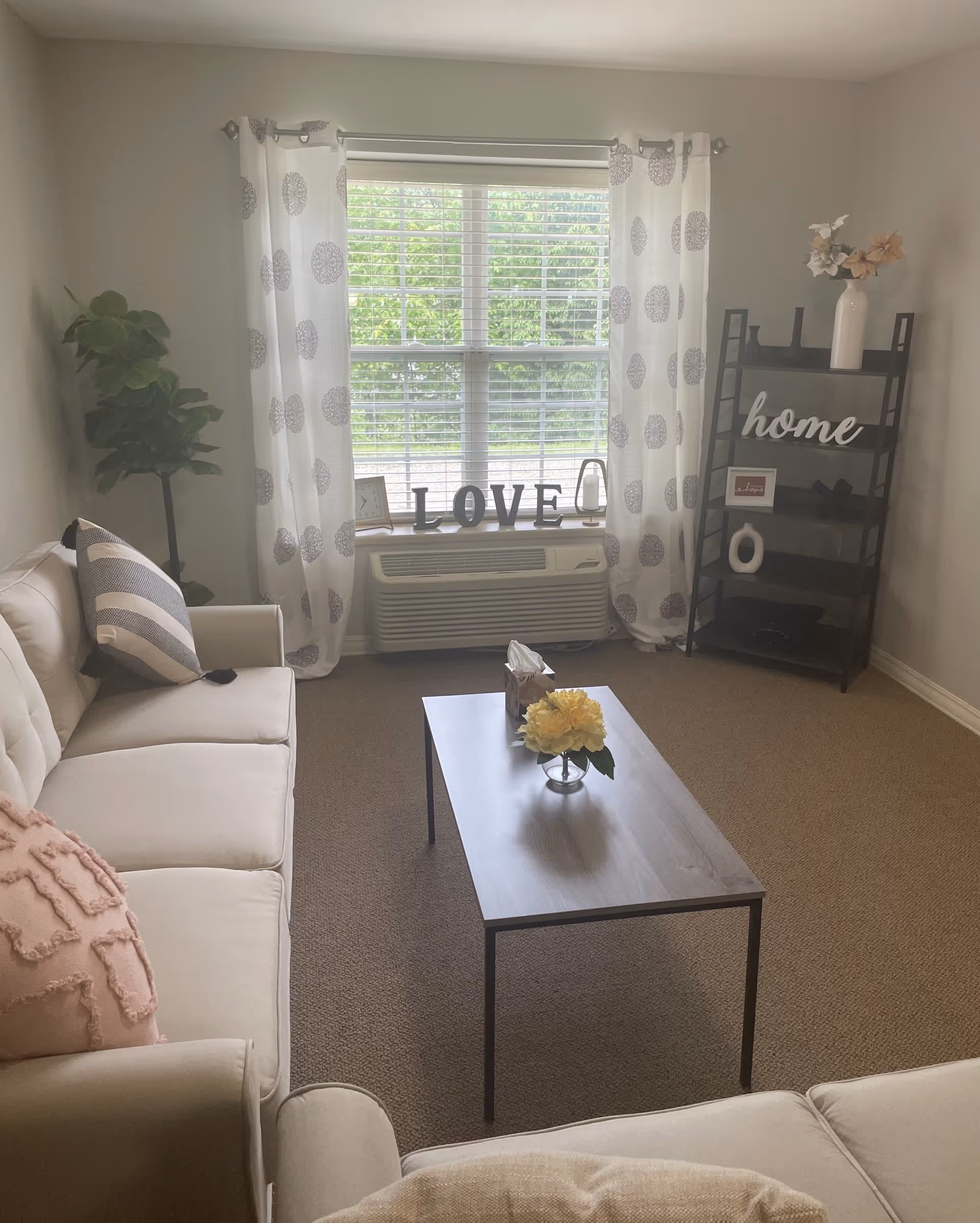 A cozy living room with beige sofas adorned with pillows, a wooden coffee table with a yellow flower centerpiece, a window with white curtains featuring circular patterns, and a black shelving unit with decorative items including a vase, a framed picture, and the word 'home'. The window sill has decorative letters spelling 'LOVE'.