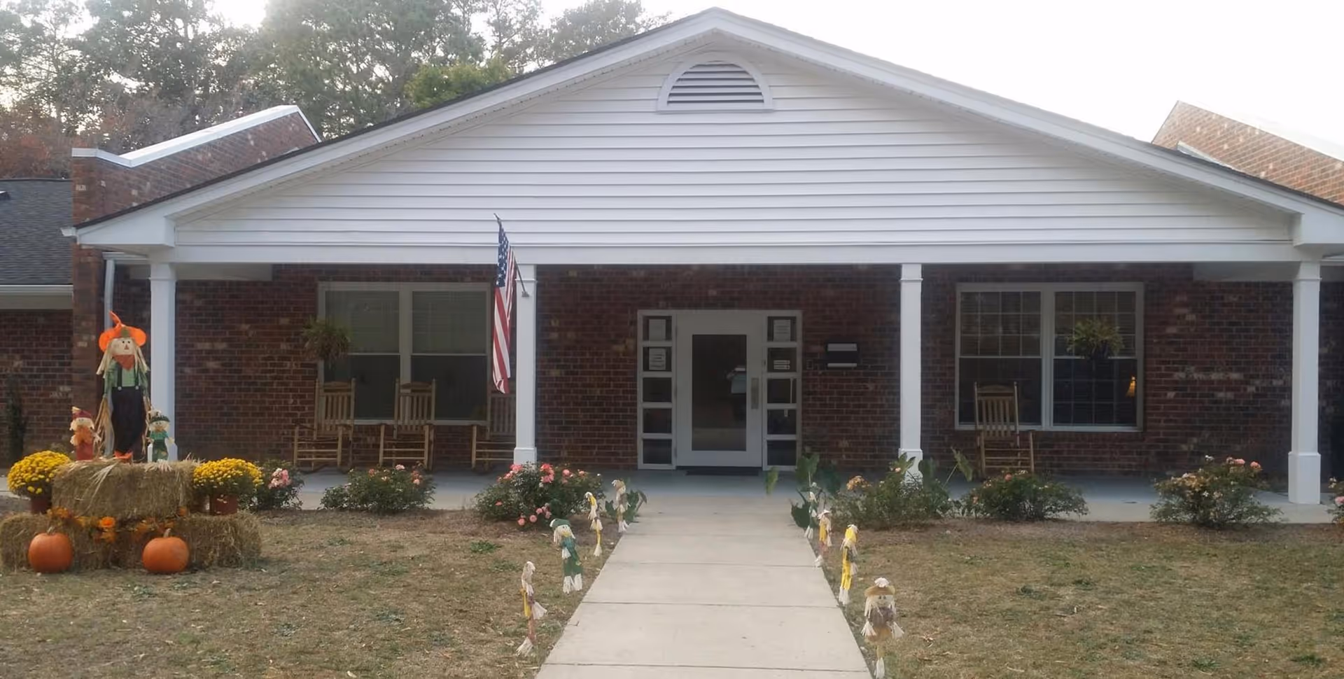 Front exterior view of a single-story brick building with a white gabled roof and a covered porch. The porch has rocking chairs and hanging plants. There is an American flag mounted on one of the porch columns. The lawn in front features fall decorations including hay bales, pumpkins, and scarecrows along the walkway leading to the entrance.