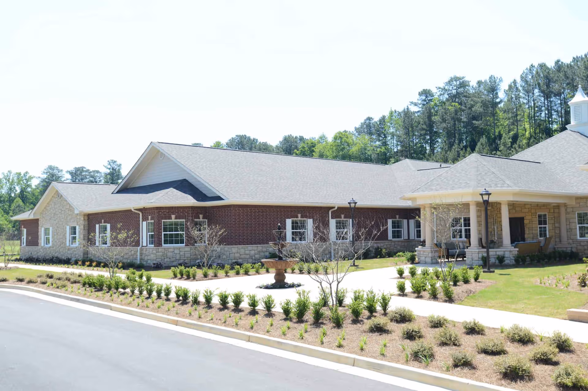 Exterior view of a single-story senior living facility building with a combination of brick and stone facade, surrounded by landscaped greenery and a paved driveway. The building has multiple windows and a covered entrance area with columns and outdoor seating. Trees and a clear sky are visible in the background.