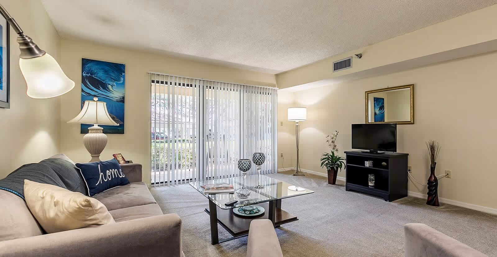 A cozy living room with a beige sofa adorned with pillows, a glass coffee table with decorative items, a TV on a black stand, a floor lamp, and a large window with vertical blinds letting in natural light. The walls are light-colored with a framed mirror and artwork.