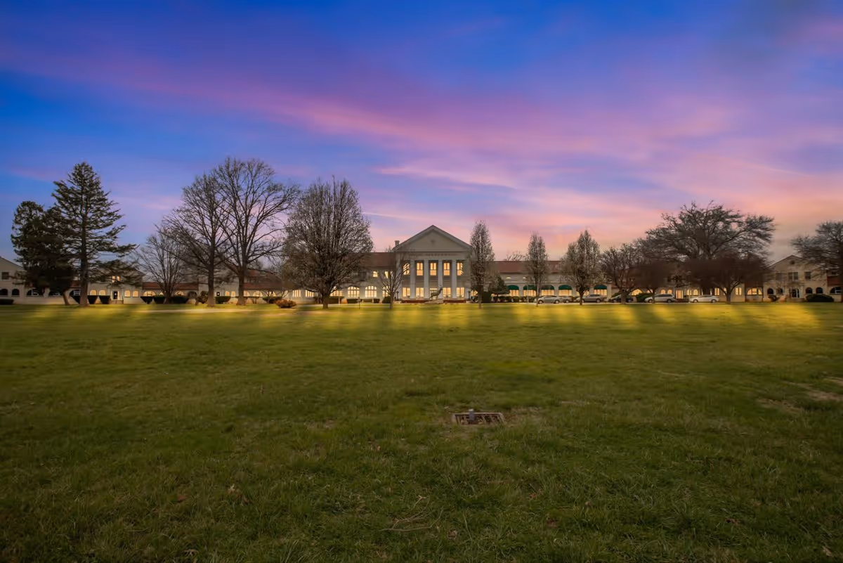 Wide view of a large senior living facility building named Spring Oak at Bedford - The Elks Home, with a well-maintained green lawn in the foreground and several leafless trees. The sky is colorful with shades of purple, pink, and blue during sunset or sunrise.