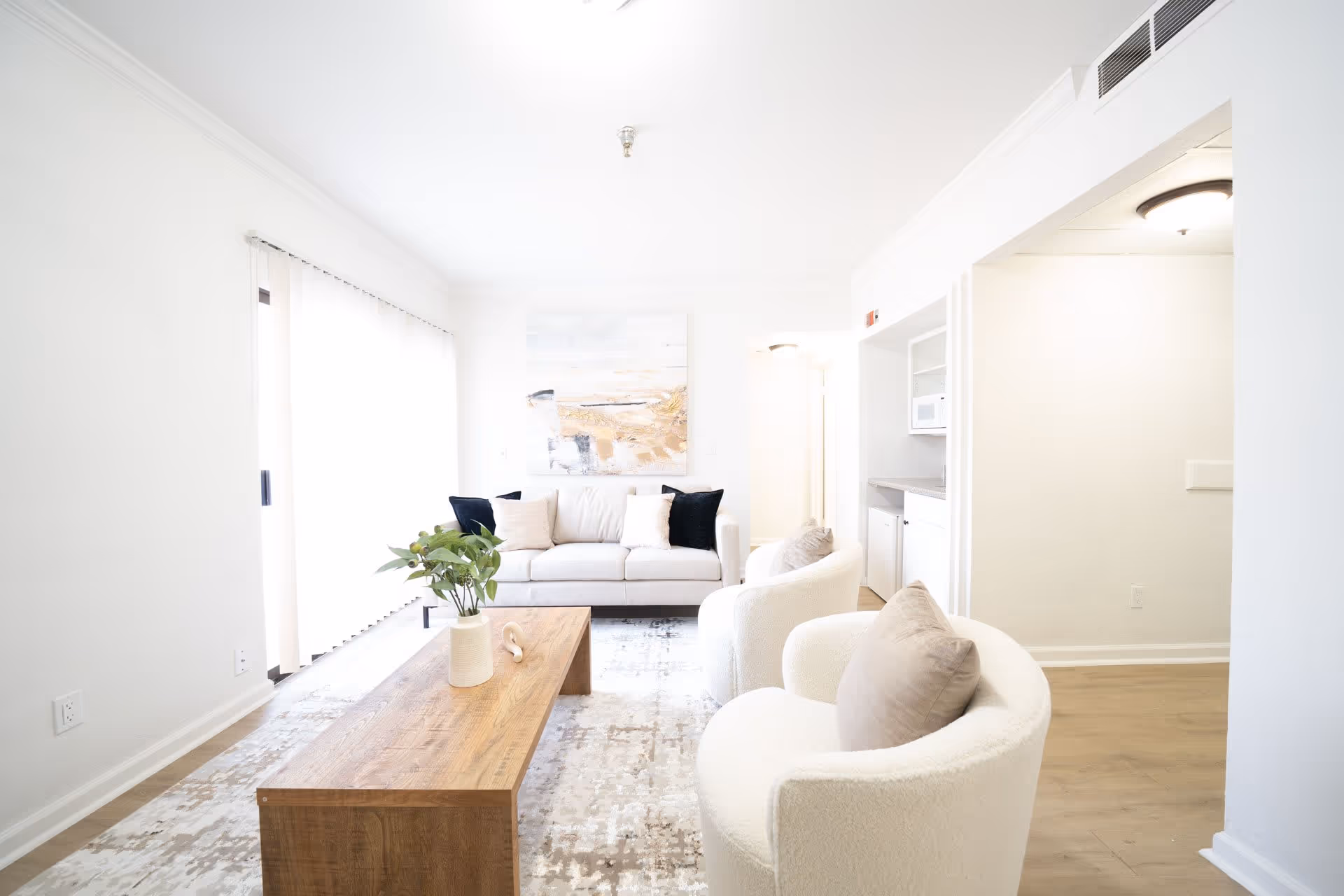 Bright and airy living room with a white sofa adorned with black and beige pillows, two white cushioned armchairs with beige pillows, a wooden coffee table with a plant in a white vase, and a large abstract painting on the wall. The room has light-colored walls, a patterned rug, and a small kitchenette area visible in the background.