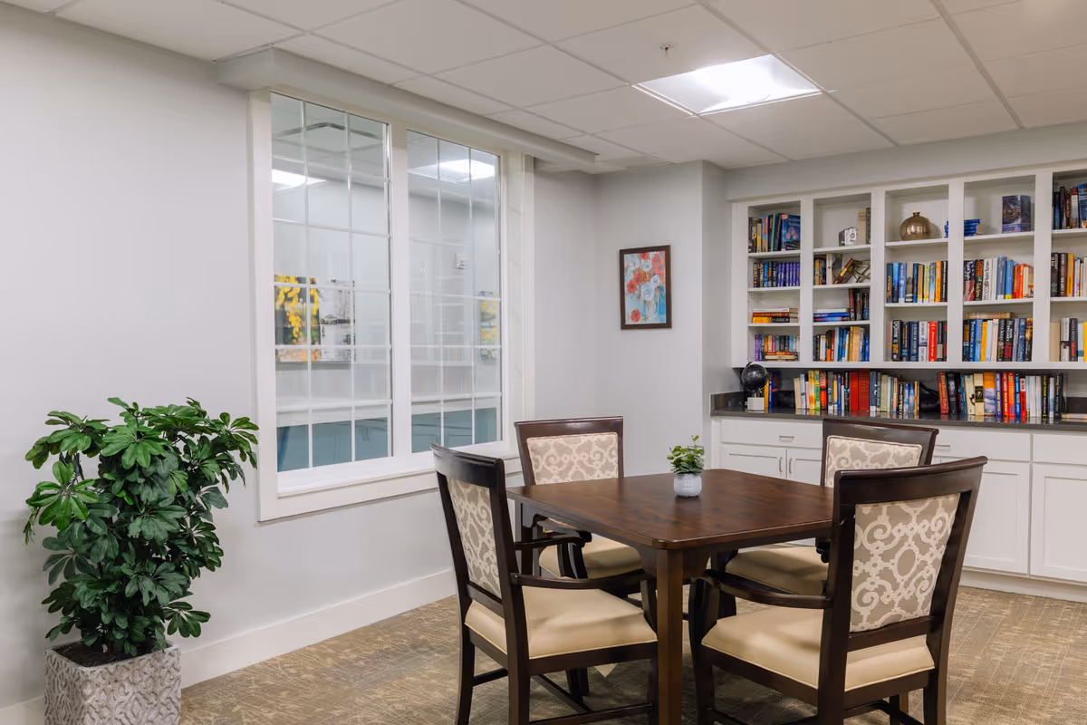 A cozy reading or meeting area with a wooden table surrounded by four cushioned chairs with patterned backs. Behind the table is a built-in bookshelf filled with books and decorative items. A small potted plant sits on the table. The room has light-colored walls, a large window with multiple panes, a framed floral painting on the wall, and a green potted plant in a decorative pot on the floor.