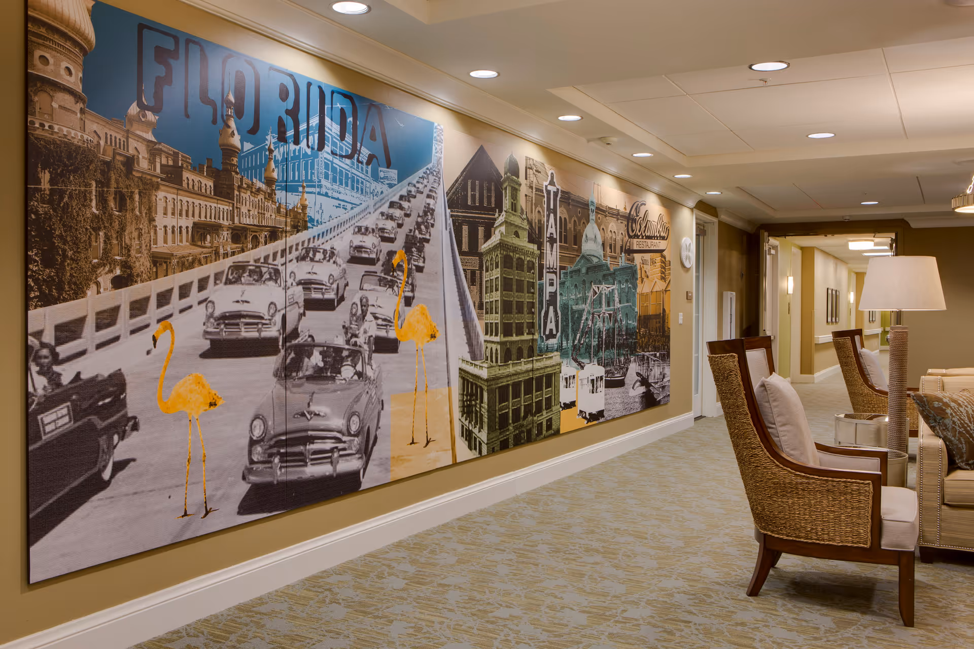 A hallway in a senior living facility with a large mural on the left wall depicting vintage Florida scenes including classic cars, flamingos, and historic buildings. On the right side, there are comfortable chairs with cushions and a floor lamp, leading to a corridor with doors and framed pictures on the walls.