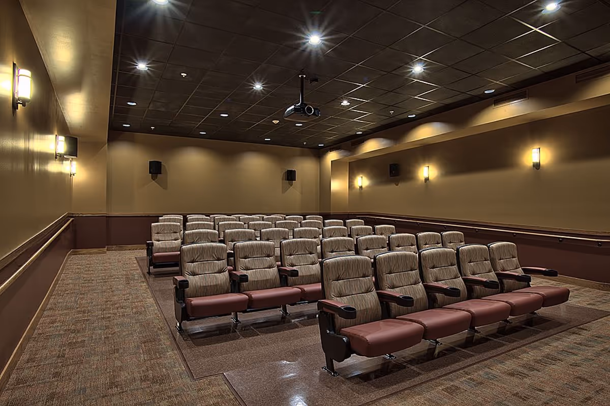 Small movie theater screening room with rows of upholstered seats, wall sconces, and a ceiling-mounted projector.