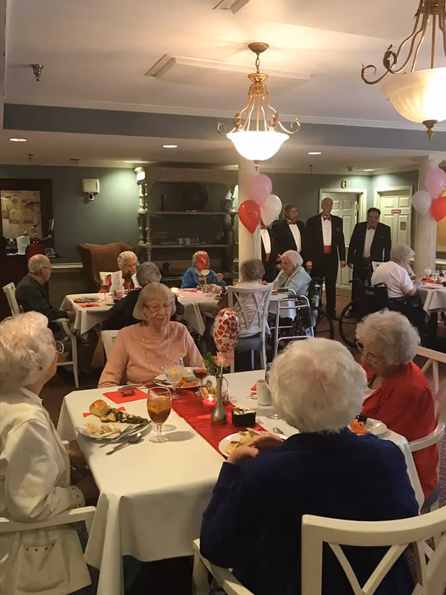 A group of elderly people seated at tables in a decorated dining room, enjoying a meal together. The tables are covered with white tablecloths and red runners, with heart-themed balloons and small floral centerpieces. In the background, four men in tuxedos stand near the wall, possibly preparing to perform or speak. The room has warm lighting and a cozy atmosphere.