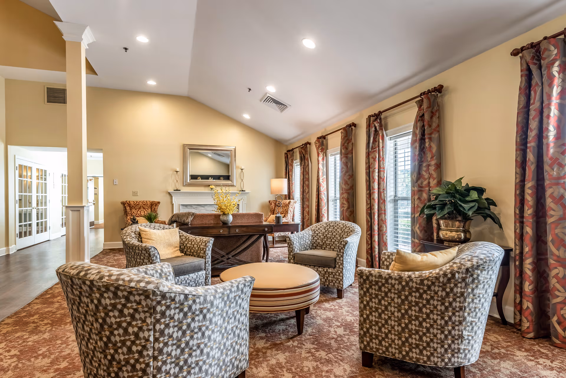 A cozy living room area in a senior living facility with four patterned armchairs arranged around a round ottoman. The room features large windows with patterned curtains, a carpeted floor, a table with a vase of yellow flowers, a fireplace with a mirror above it, and warm lighting.
