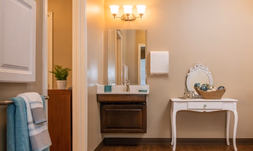 Vanity area with a wall-mounted sink and mirror, towel rack on the left, and a small white dressing table with decorative items on the right.