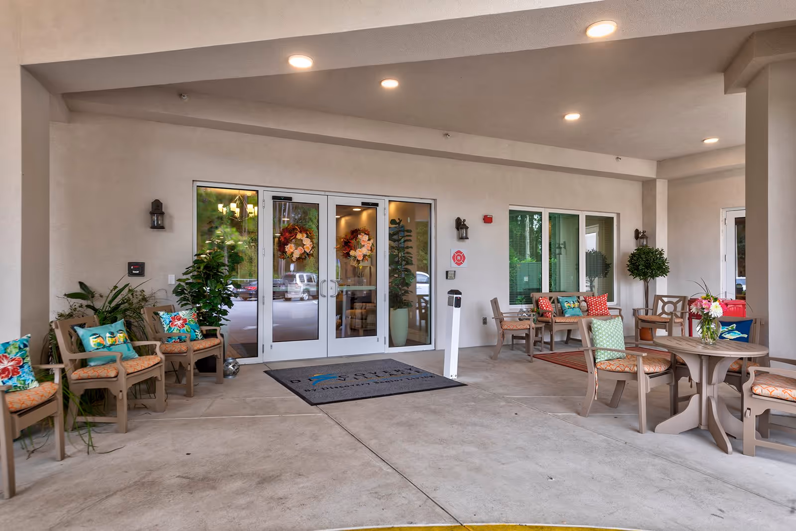 Covered entrance area of a senior living facility with multiple chairs and tables arranged for seating. The chairs have colorful cushions and there are potted plants around. Double glass doors with floral wreaths lead inside the building.