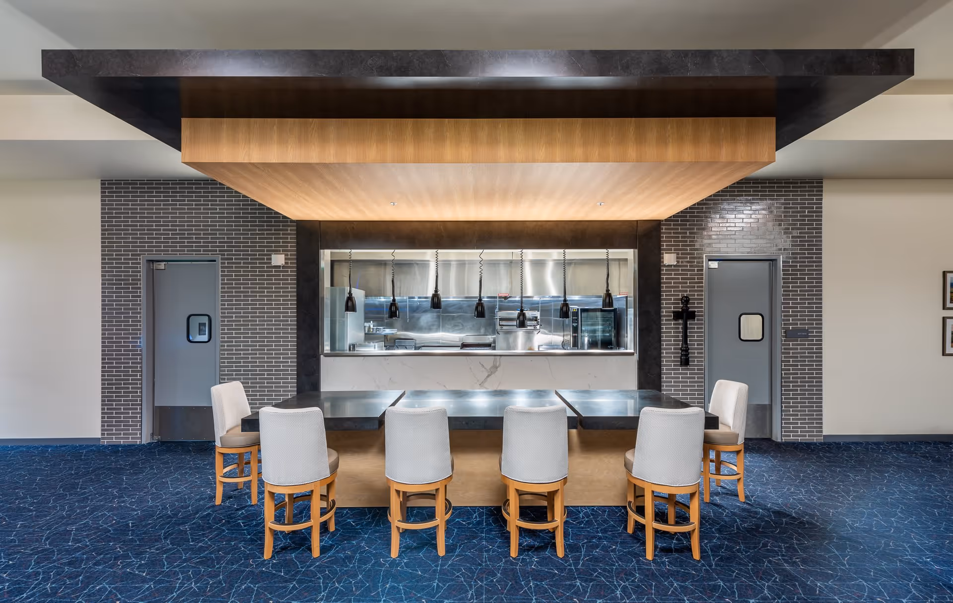 A modern dining area with a long table surrounded by six cushioned chairs with wooden legs. Behind the table is a kitchen pass-through window with hanging pendant lights and a visible stainless steel kitchen. The walls are decorated with gray brick tiles and two gray doors on either side of the kitchen window. The ceiling features a wooden panel with recessed lighting above the table, and the floor is covered with a blue patterned carpet.