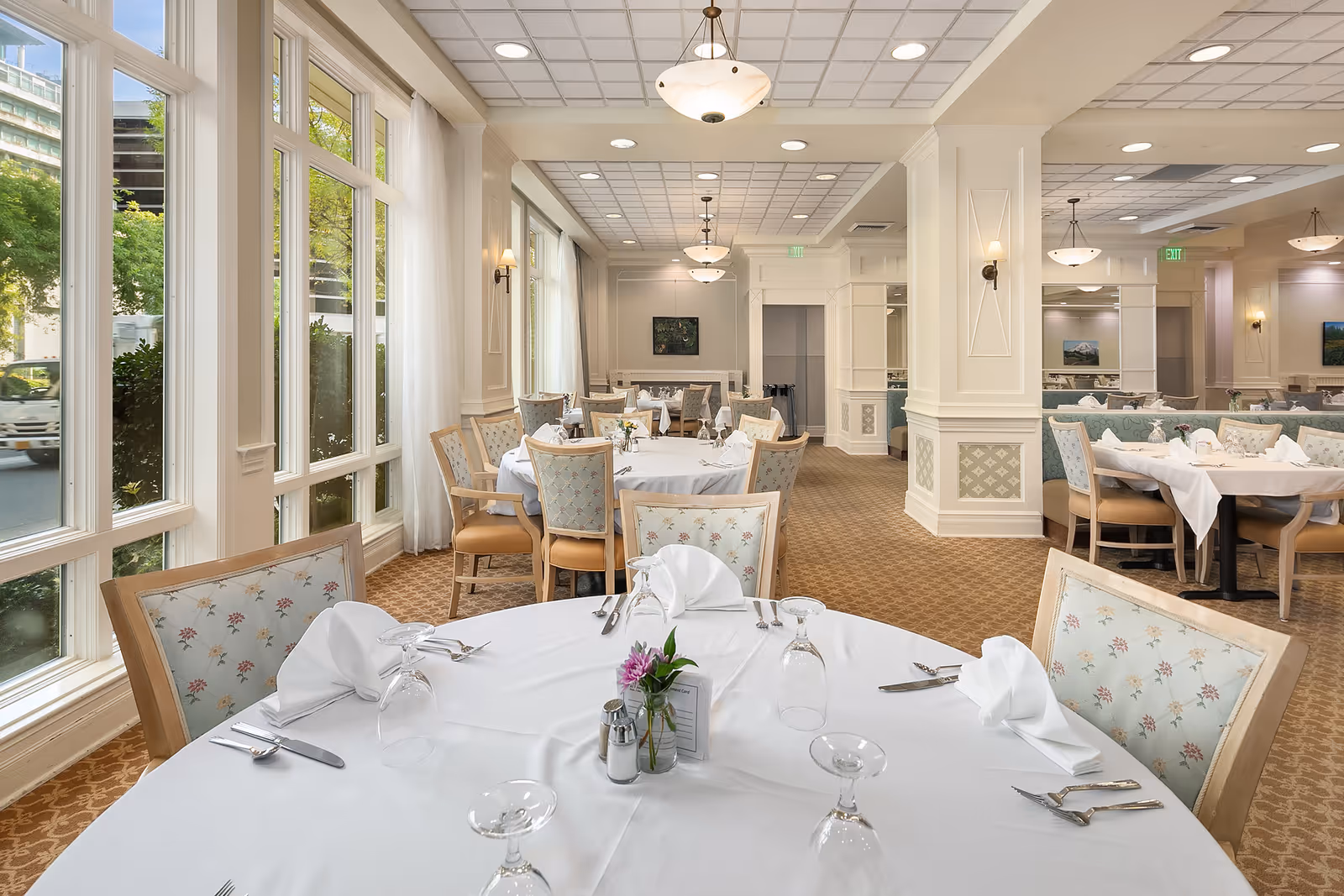 A bright and elegant dining room with round tables covered in white tablecloths, set with folded napkins, glassware, and silverware. The room features large windows with sheer curtains allowing natural light to fill the space, patterned carpet, and upholstered chairs with floral designs. Soft ceiling lights and wall sconces provide additional lighting.