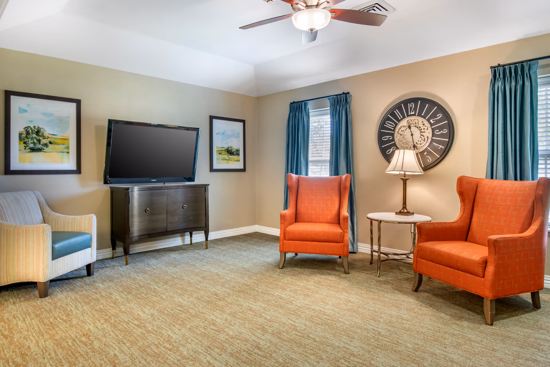 A cozy living room area with two orange wingback chairs and one striped armchair. There is a small round table with a lamp between the orange chairs. A large wall clock hangs above the table. Two windows with blue curtains let in natural light. A flat-screen TV is mounted on a dark wooden cabinet, flanked by two framed landscape paintings. The room has beige walls, a ceiling fan with a light, and beige carpeted floor.