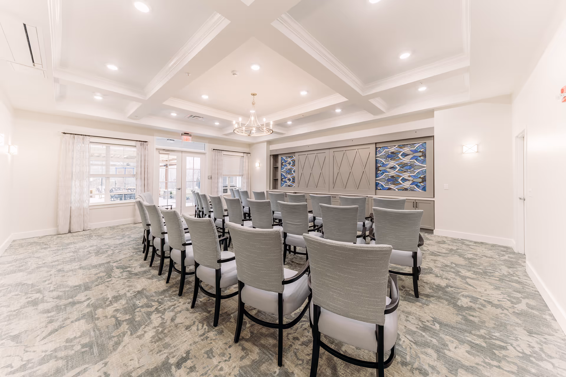 A bright, modern meeting or conference room with rows of gray upholstered chairs arranged facing a wall with decorative panels featuring blue and gray patterns. The room has a coffered ceiling with recessed lighting and a central chandelier. Large windows with sheer curtains and a glass door provide natural light and a view of an outdoor patio area.