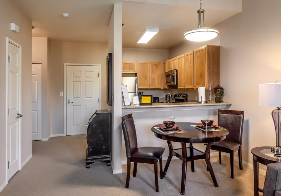Open-concept senior apartment kitchen and dining area with wooden cabinets, a countertop bar, and a round dining table set for two.