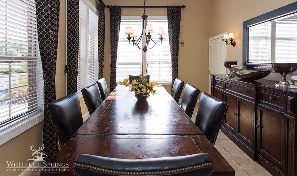 A dining room with a long wooden table surrounded by black leather chairs. The table has a floral centerpiece. The room has large windows with patterned curtains, a chandelier hanging above the table, and a dark wooden sideboard with decorative bowls and a large mirror above it.