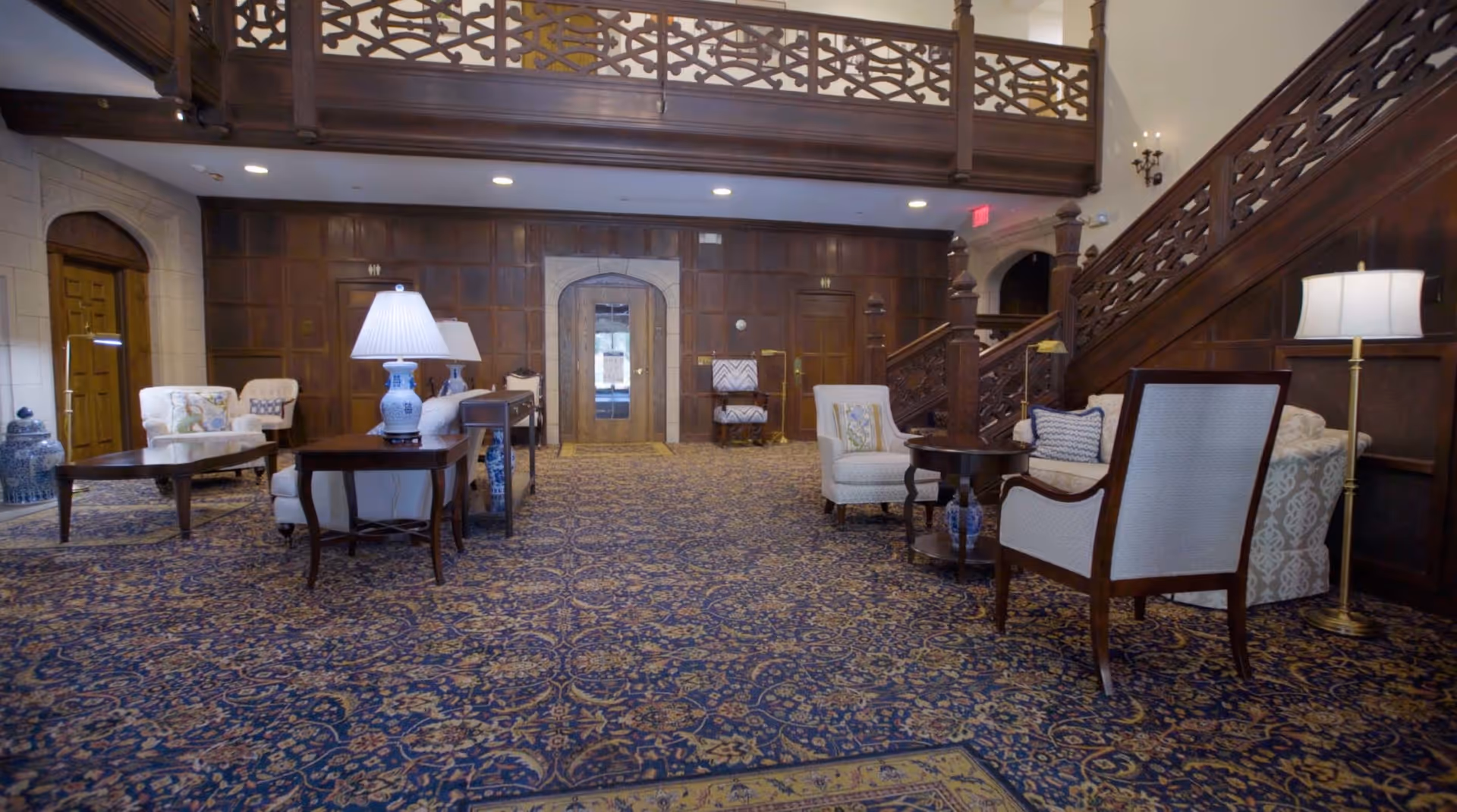 A spacious, elegant lounge area with patterned carpet, wooden paneling on the walls, and a wooden staircase with decorative railing. The room is furnished with several upholstered armchairs, side tables with lamps, and decorative vases. There is a door with a glass panel at the far end of the room.