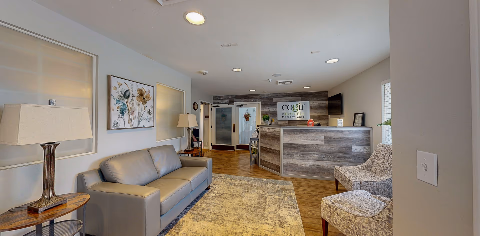 Bright reception lounge with a gray sofa, patterned chairs, lamps, rug, and a wood-paneled reception desk bearing a Cogir of Bothell Memory Care sign.