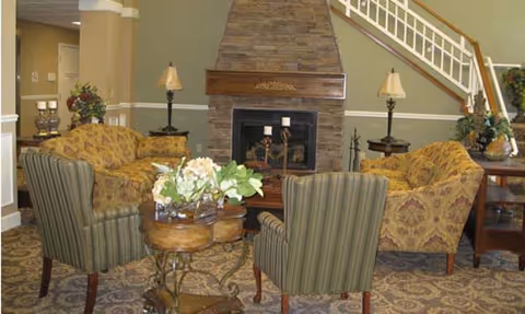 A cozy living room area with a stone fireplace in the center, flanked by two patterned sofas and two striped armchairs. There are two side tables with lamps on either side of the fireplace, a small round table with a flower arrangement in the middle, and a staircase with white railings in the background.