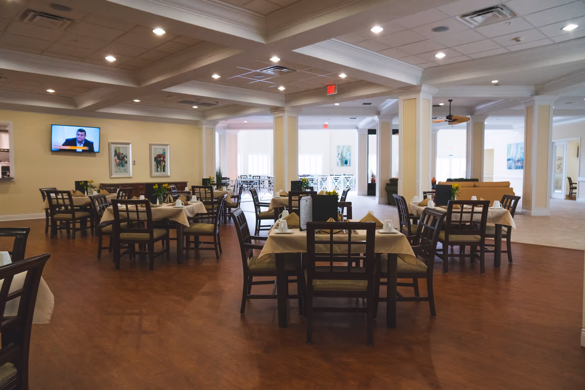 A spacious dining room in a senior living facility with multiple tables covered with beige tablecloths, each set with cups and napkins. The room has wooden floors, cream-colored walls, and ceiling lights. A television is mounted on one wall, and there are framed artworks and plants decorating the space.