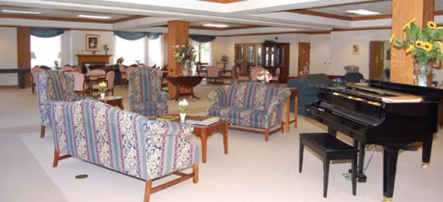 A spacious living room area with floral patterned sofas and armchairs arranged around small wooden tables. A black grand piano with a matching bench is positioned on the right side. The room features wooden columns, a carpeted floor, and large windows with curtains allowing natural light to enter. There are flower arrangements on tables and a wooden cabinet in the background.