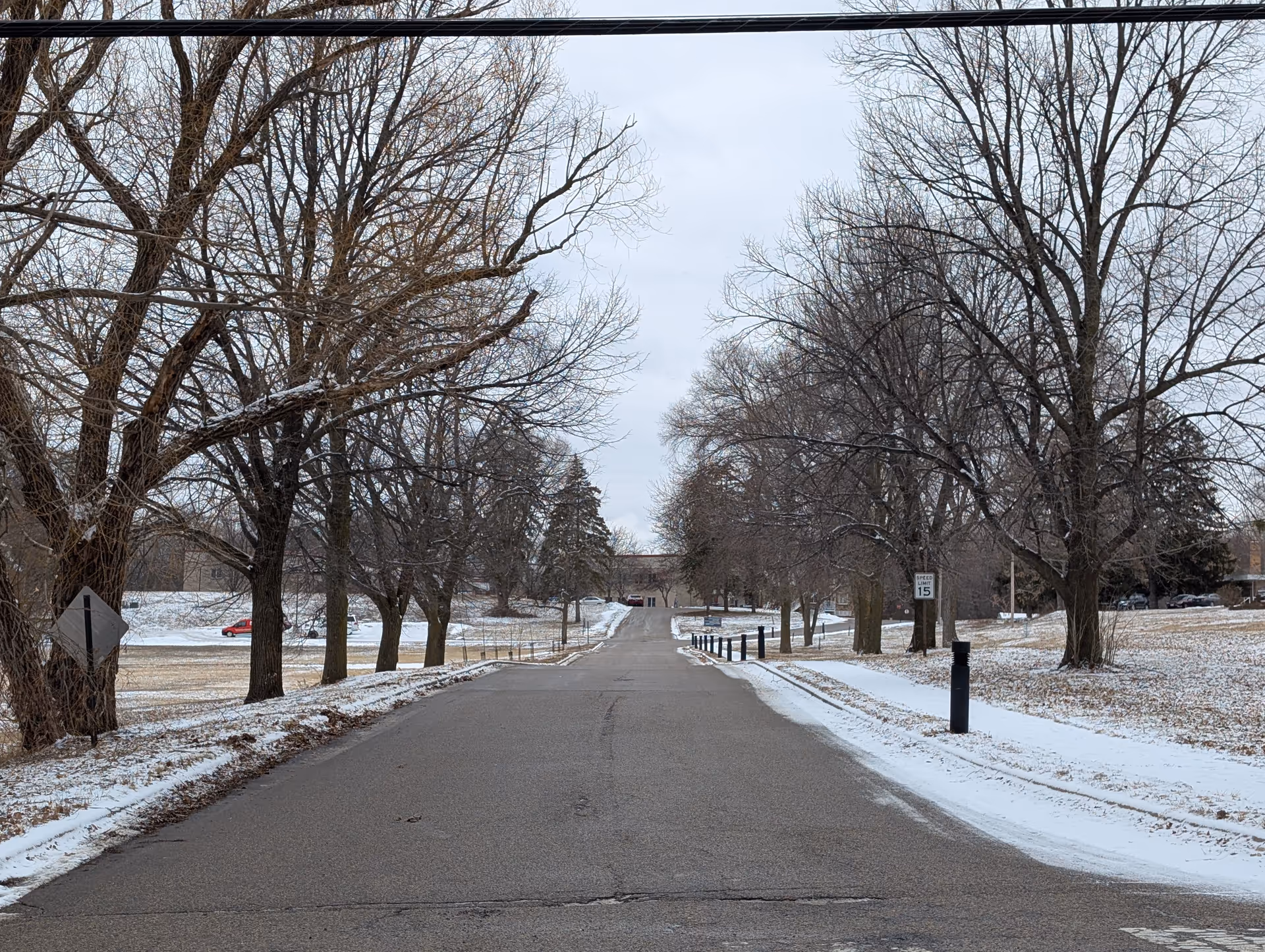 A paved road lined with leafless trees on both sides during winter, with patches of snow on the ground and a speed limit sign showing 15 mph. A building is visible in the distance at the end of the road.